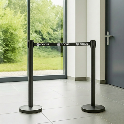 Two black Cocus retractable belt and post barriers with a black belt, displayed in a modern lobby with a tiled floor and large windows showing green trees outside.
